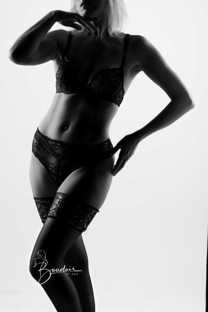 Venisis debout en lingerie et bas, une main près de l’épaule et l’autre sur la hanche, en noir et blanc.