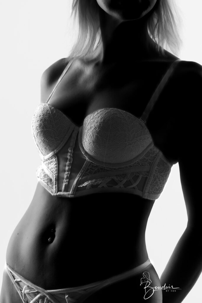 Venisis debout en corset clair, la silhouette éclairée de côté sur fond sombre, en noir et blanc.